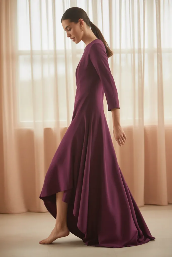 A medium full-length photo of a woman walking through a sunlit hallway wearing a plus size night dress for women in deep teal blue. Features a curved high-low hemline, three-quarter length sleeves, and a soft scoop neck. Warm natural lighting, clean minimalist home environment, graceful fluid motion, standard perspective angle.